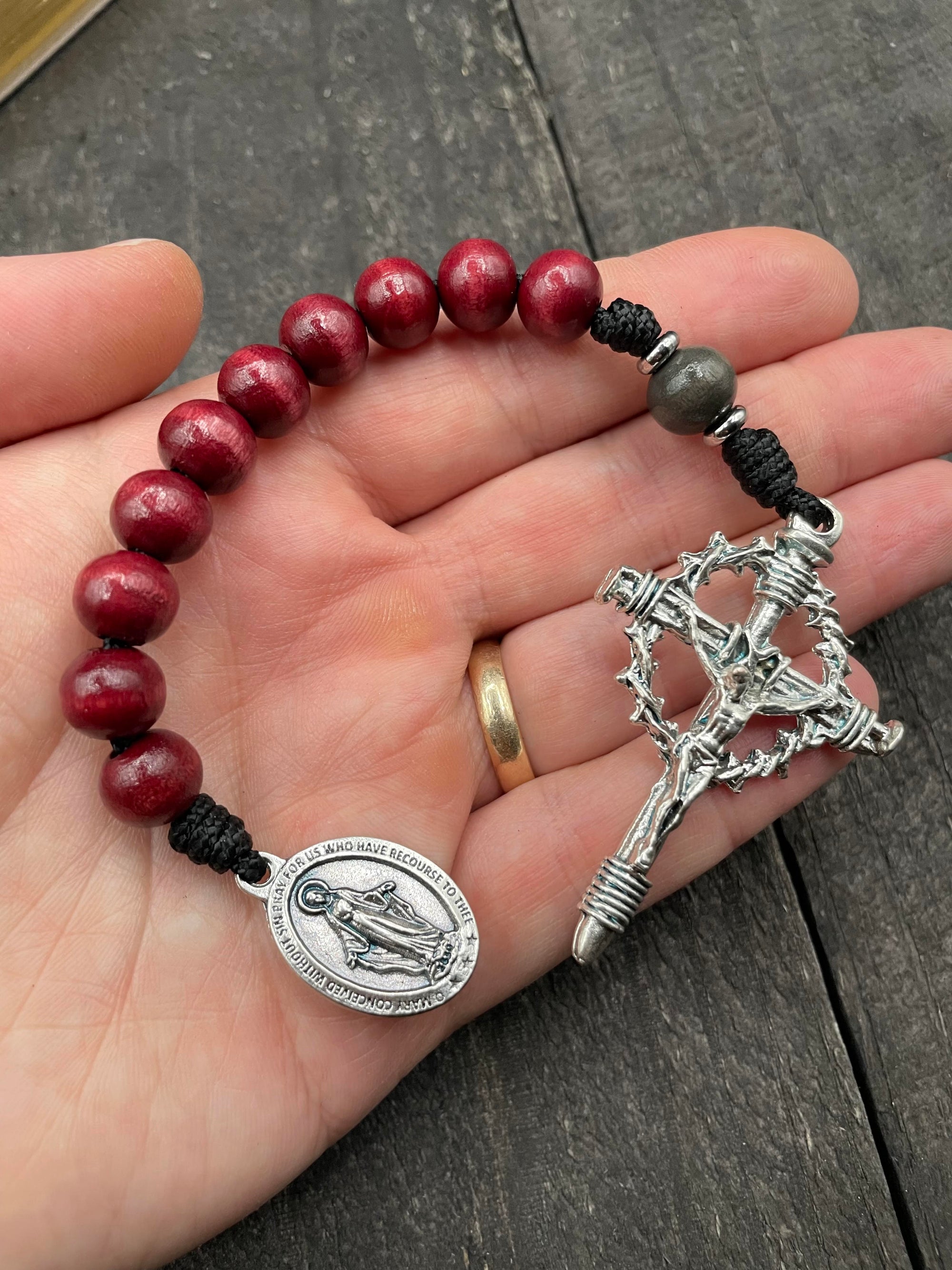 Hand holding a rosary bracelet featuring red wooden beads, a silver crucifix with a thorn crown design, and a silver medallion depicting a religious figure.