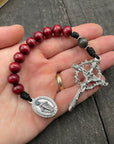 Hand holding a rosary bracelet featuring red wooden beads, a silver crucifix with a thorn crown design, and a silver medallion depicting a religious figure.