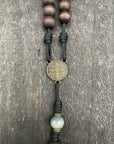 Close-up of a rosary with wooden beads, a Saint Benedict medal, intricate black cord knots, and a larger central bead, displayed on a textured gray surface, symbolizing spiritual devotion and protection.