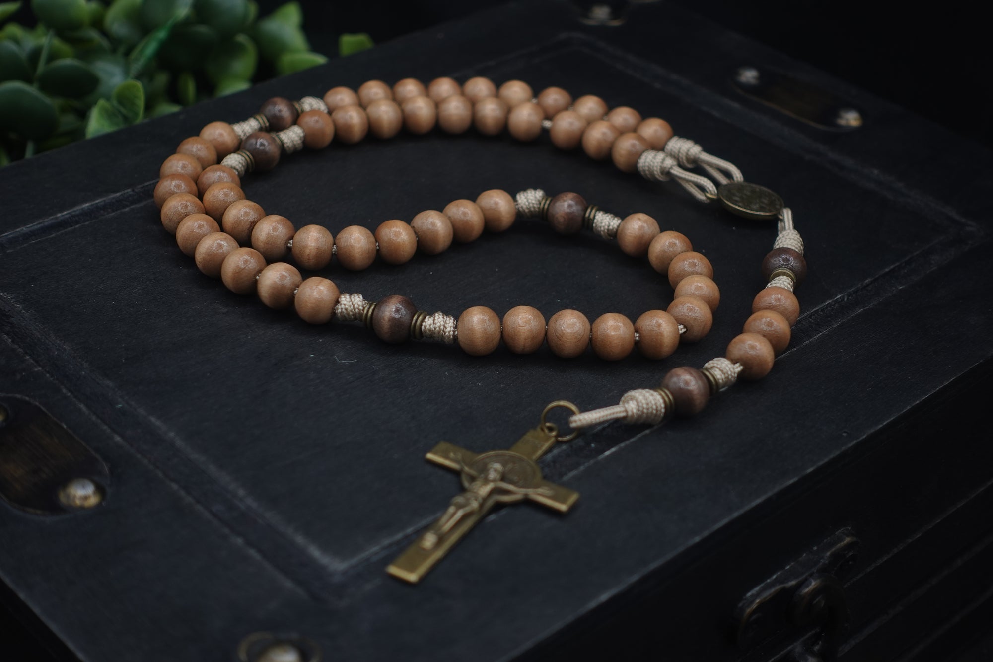 paracord rosary with tan and gray-brown wooden beads and bronze St. Benedict crucifix resting on black box