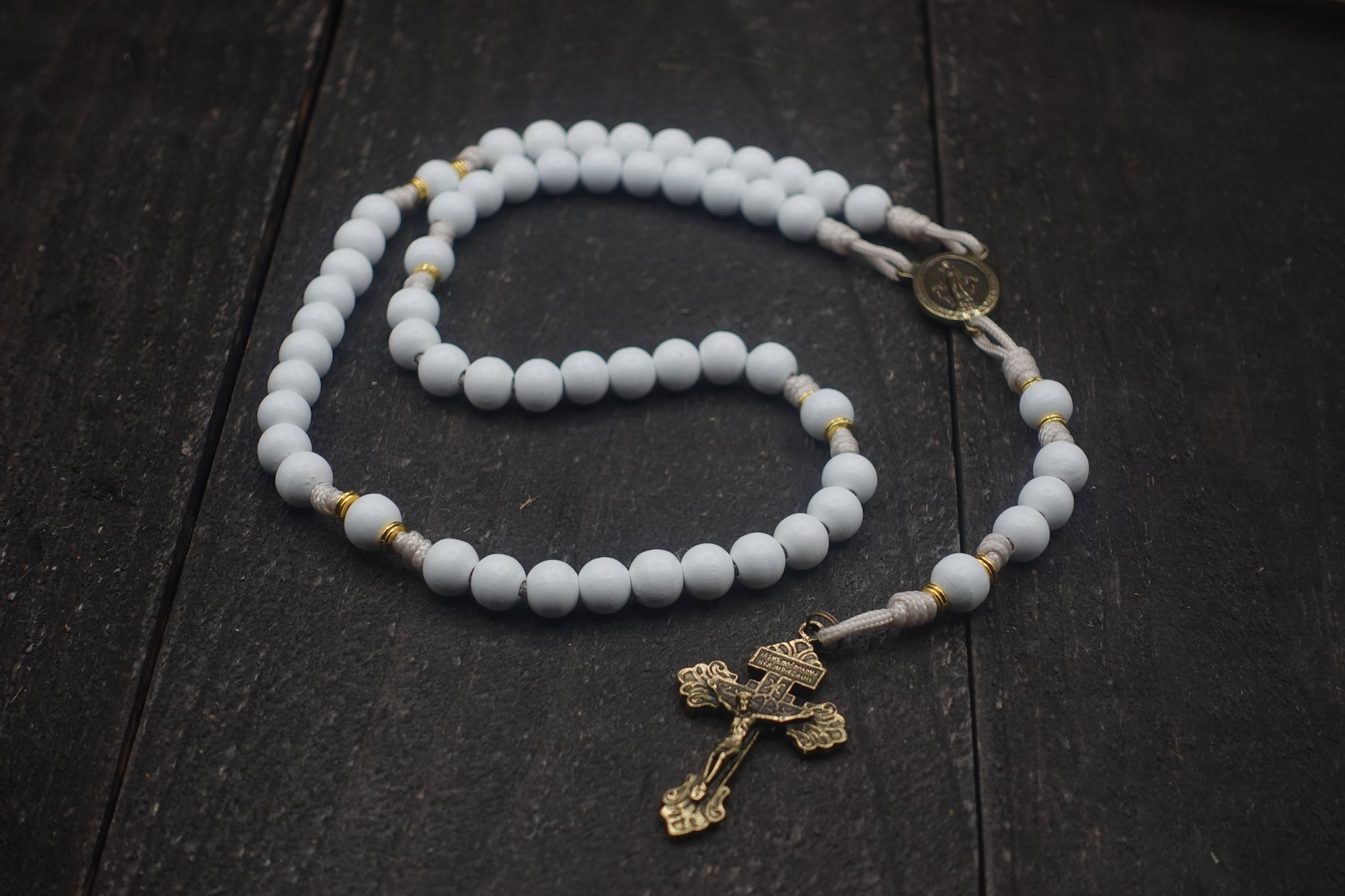 Handcrafted rosary featuring white wood beads, bronze crucifix, and gold spacer accents on cream paracord