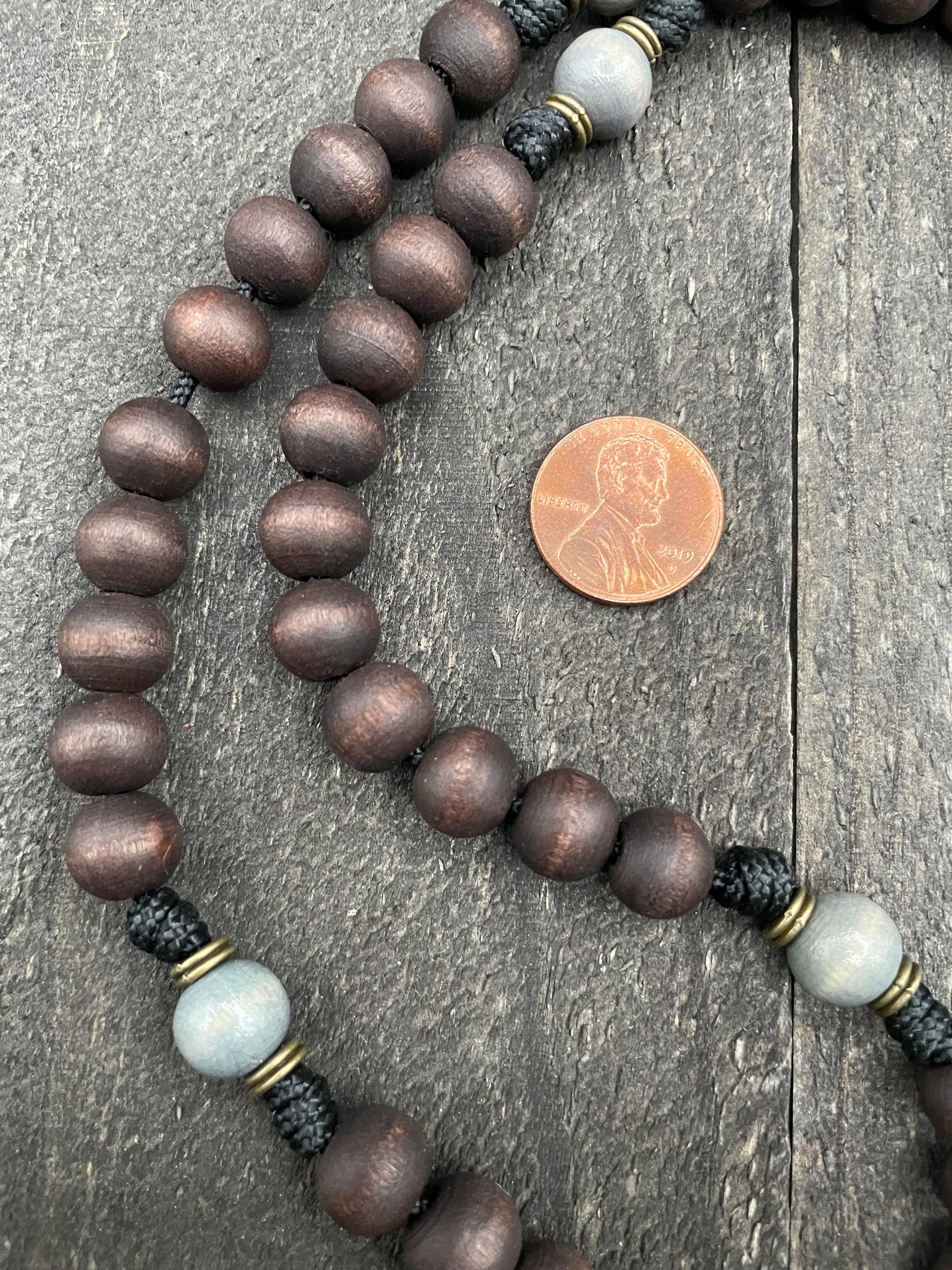 Close-up of a dark wooden bead rosary with a penny for size reference, displayed on a textured wooden surface, highlighting its craftsmanship and material quality.