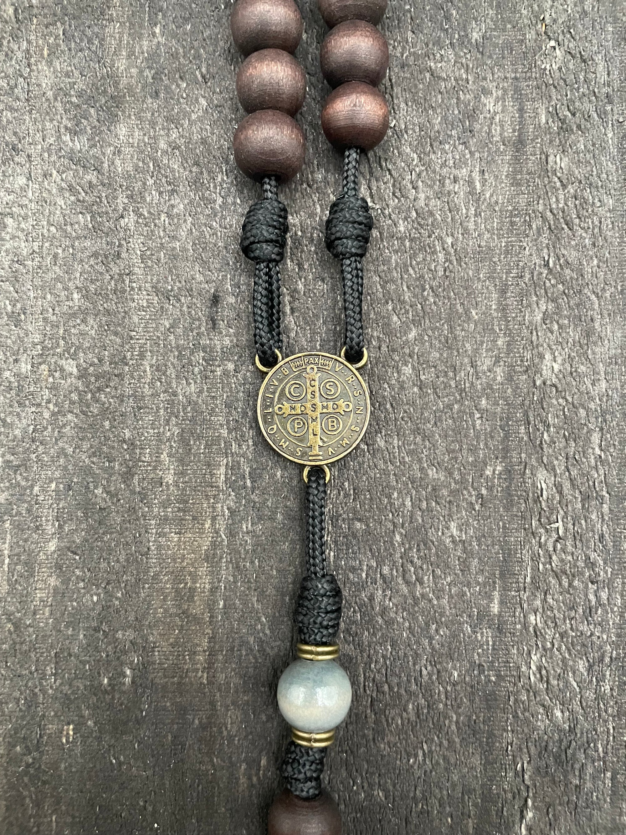 Close-up of a rosary with wooden beads, a Saint Benedict medal, intricate black cord knots, and a larger central bead, displayed on a textured gray surface, symbolizing spiritual devotion and protection.